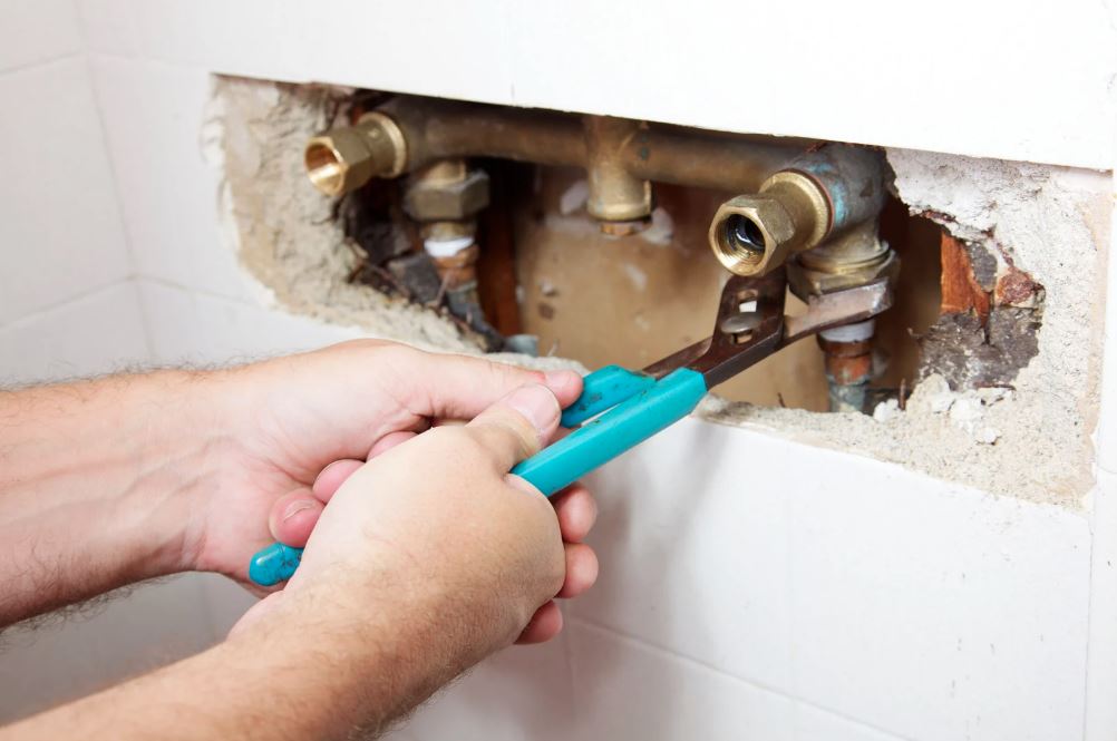 Plumber using a wrench to repair exposed water pipes inside a wall.